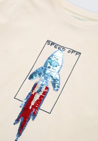 Cremfarbener T-Shirt mit einem Raketenmotiv aus blauen, roten und silbernen Pailletten, mit dem Text "SPEED OFF" darüber in Schwarz.