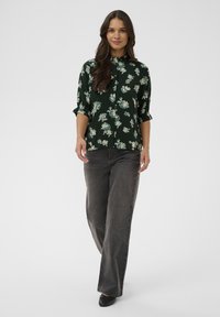 Tamsiai žalia gėlių rašto marškinėliai su suvirintomis rankovėmis, derinami su laisvais tamsiai pilkais džinsais ir juodais batais.