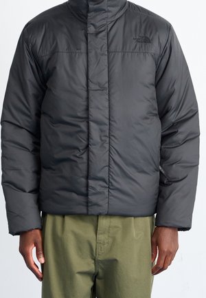 Zwart gewatteerd North Face-jas gedragen door een persoon met olijfgroene broek, staand tegen een effen lichte achtergrond.