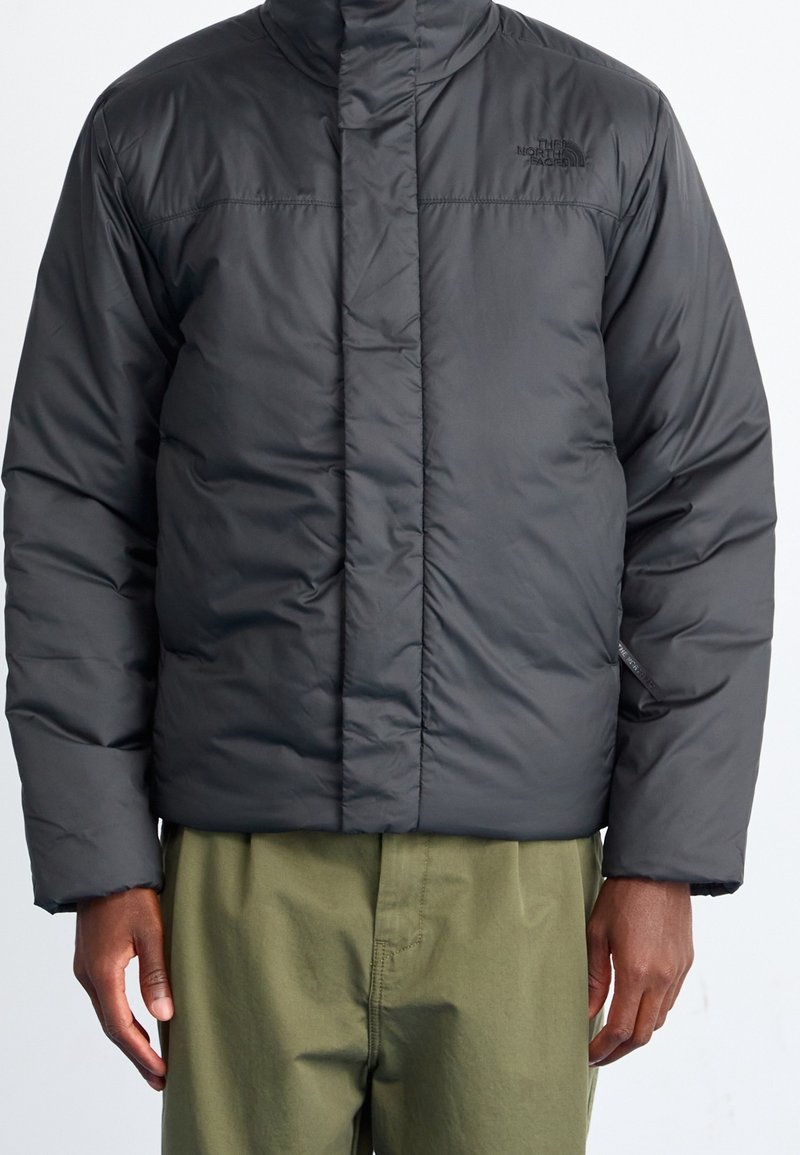 Veste matelassée noire de The North Face portée par une personne en pantalon vert olive, debout devant un fond clair uni.