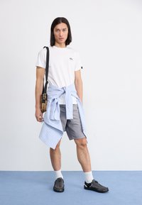 Hvit t-skjorte med liten logo, grå shorts, lys blå stripet skjorte knyttet rundt livet, svarte sko og hvite sokker. Enkelt design og uformell stil.