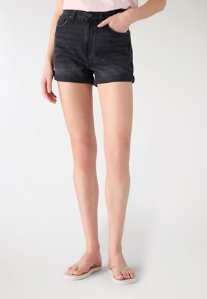 Donna che indossa pantaloncini di denim neri con orlo risvoltato, top rosa chiaro e infradito beige, in piedi con una mano in tasca su sfondo semplice.