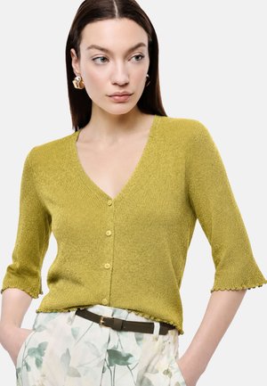 Donna che indossa un cardigan giallo senape texturizzato con bottoni, maniche a tre quarti e pantaloni con motivo floreale e cintura nera.