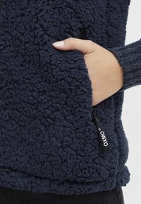 Ruka v kapse námorně modré fleecové bundy s texturou, se zipem označeným značkou "OXMO".