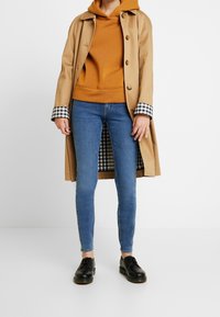 Beige trench coat with buttons, paired with an orange hoodie. Blue slim jeans and black lace-up shoes complete the outfit. Checkered cuff detail.