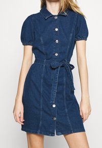 Kvinna klädd i en kortärmad, mörkblå jeansklänning med framknapningar och ett midjeband i form av en båge.