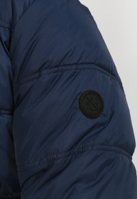 Rukáv granátově modré prošívané bundy s elastickými manžetami. Na rameni je výrazně umístěna kulatá černá nášivka s logem.