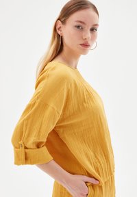 Mosterdgele, gestructureerde longsleeve top met een ronde halslijn en omgeslagen mouwen, voorzien van subtiele verticale plooien en een relaxte pasvorm.
