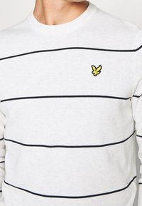 Maglione bianco con strisce blu navy, collo a coste tondo e piccolo logo di aquila gialla sul petto sinistro. Texture morbida, design casual.