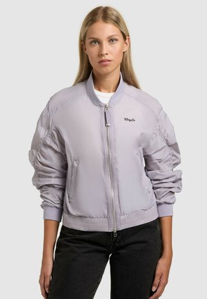 Młoda kobieta ubrana w jasnofioletową kurtkę bomber z suwakiem i marszczonymi rękawami oraz czarne spodnie, stojąca na jednolitym tle.