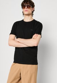 Čierne tričko s krátkym rukávom a klasickým výstrihom, na hrudi s jemným logom "BOSS". Model má na sebe hnedé slnečné okuliare a béžové nohavice.