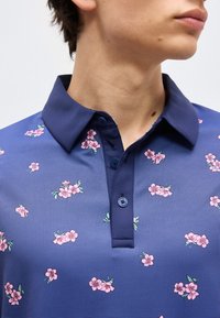 Κοντινό πλάνο ενός ατόμου που φοράει μπλε-σκούρο πόλο πουκάμισο με μικρό ροζ ανθικό μοτίβο και τρία μπλε κουμπιά στη λαιμόκοψη.
