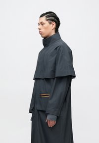 Tmavomodrý dáždnikový kabát s vysokým golierom a viacvrstvovým plášťom. Má zipsovou kapsu s oranžovo-žltým pruhom. Hladká textúra.