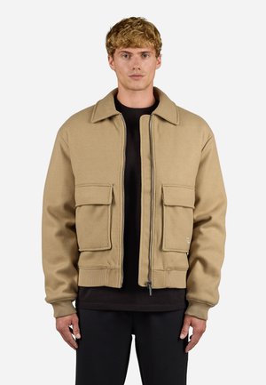 Beige Bomberjacke mit Reißverschluss, zwei großen Fronttaschen, gerippten Bündchen und einem flachen Kragen, aus weichem, strukturiertem Stoff hergestellt.