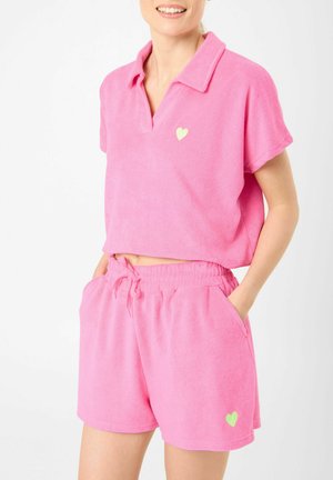 Femme portant un ensemble short en éponge rose assorti composé d’une chemise à col et d’un short à taille élastique, chacun avec un petit détail en forme de cœur.
