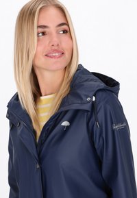 Chaqueta de lluvia de color navy, hecha de material impermeable y con capucha. Presenta un logo de paraguas plateado y detalles de cremallera. Camiseta amarilla a rayas visible debajo.