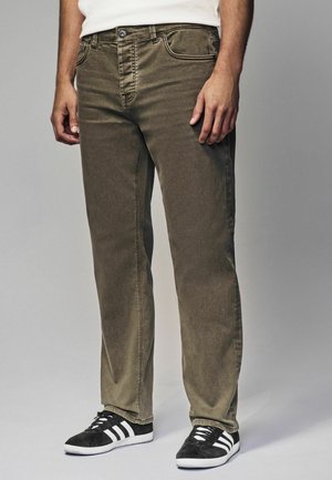 Jeans en denim vert olive avec une coupe droite, cinq poches et des coutures contrastées. Associés à des baskets noires avec des rayures blanches.