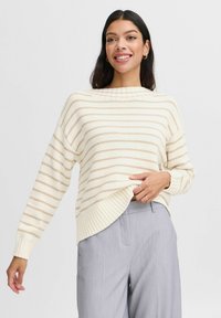 Cremestrickpullover mit beigen horizontalen Streifen, geripptem Saum und Bündchen, kombiniert mit hellgrauen Hosen. Weiche Stricktextur.
