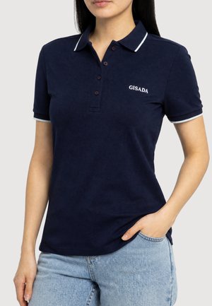 Femme portant un polo bleu marine à manches courtes avec bordure blanche et logo "GISADA", associé à un jean bleu clair, main dans la poche.