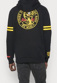 Felpa con cappuccio nera caratterizzata da un grande logo giallo di Cobra Kai sulla schiena, strisce gialle sulle maniche e testo rosso sulla parte inferiore della manica.