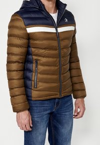 Homme portant une veste matelassée marron et marine avec une bande blanche traversant la poitrine, fermée par une fermeture éclair, associée à un jean bleu.