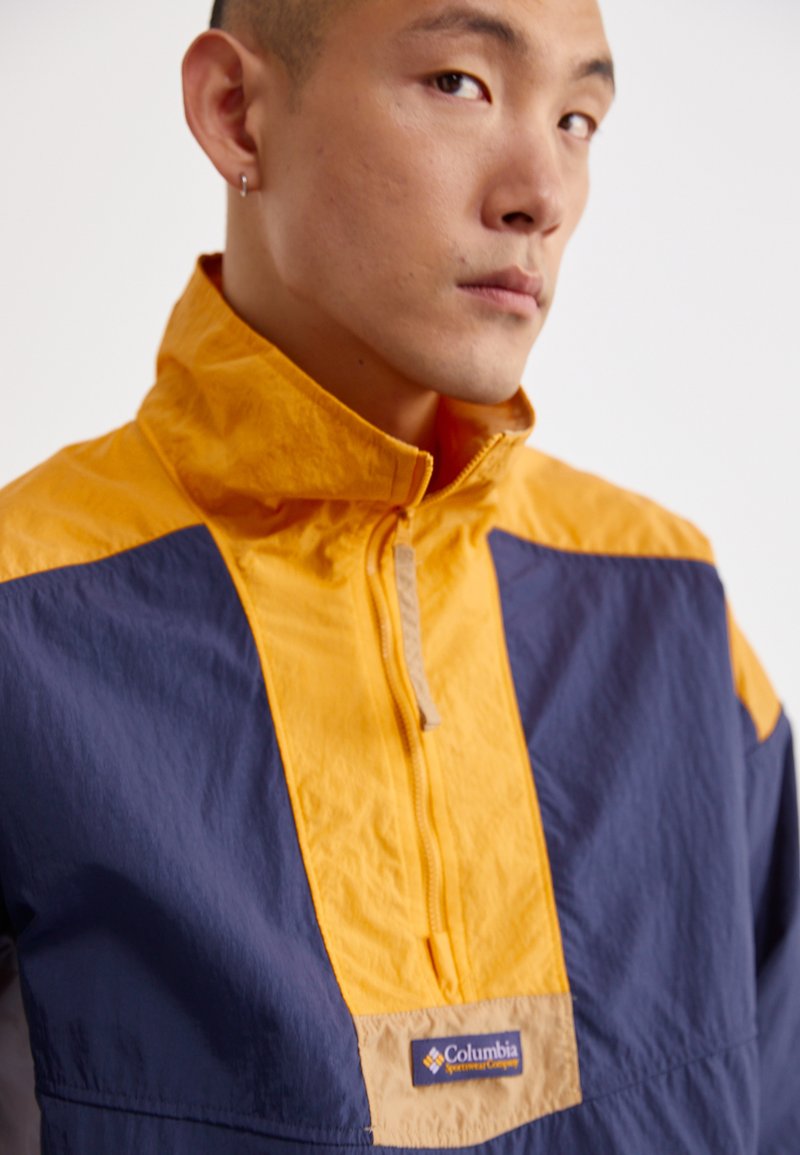 Chaqueta colorida con diseño de media cremallera, con segmentos en amarillo y azul marino. Hecha de tela ligera, incluye un logo de Columbia en la parte frontal.