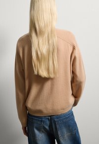 Maglione beige in lana con scollo a coste e polsini, abbinato a jeans in denim blu con tasche posteriori. Tessuto liscio e vestibilità comoda.