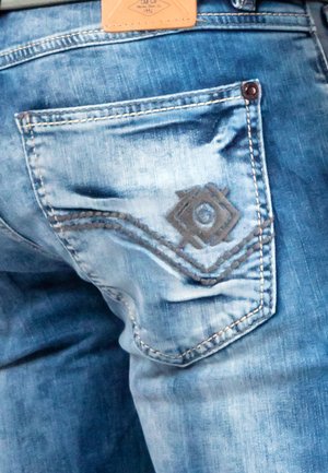 Jean en denim bleu clair avec une poche arrière ornée d'un patch marron décoratif et de détails cousus en motifs ondulés.