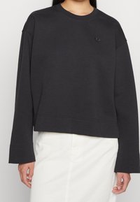 Black cropped sweatshirt with long sleeves, ribbed neckline, and tonal embroidered logo on chest. Smooth fabric with a relaxed fit.