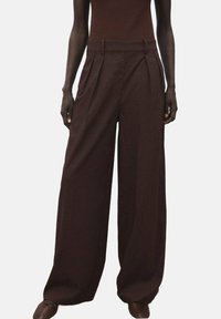 Pantalons larges en tissu côtelé marron foncé, avec des plis à la taille et une coupe décontractée. Texture lisse avec des lignes verticales subtiles.