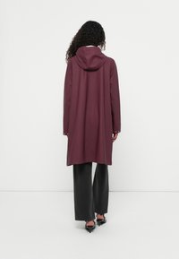 Trench coat longo de cor bordeaux com capuz, apresentando mangas longas e um corte descontraído. O material parece suave com um acabamento mate.