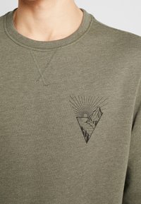 Sudadera verde oliva con acabado texturizado, cuello acanalado y una escena de montaña en negro impresa con rayos de sol en el lado izquierdo del pecho.