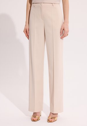 Kobieta nosząca beżowe, szerokie spodnie z wysokim stanem z przodu z fałdami, zestawione ze złotymi sandałami na wysokim obcasie z odkrytymi palcami.