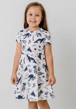 Young girl smiling, wearing a white dress with blue marine animals like octopus, whales, jellyfish, and fish printed on it.