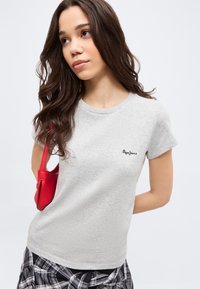 Νέα γυναίκα με μακριά σκούρα μαλλιά που φοράει γκρι ανοιχτό T-shirt Pepe Jeans, καρό πουκάμισο μαύρο-άσπρο δεμένο στη μέση και κόκκινη τσάντα ώμου.