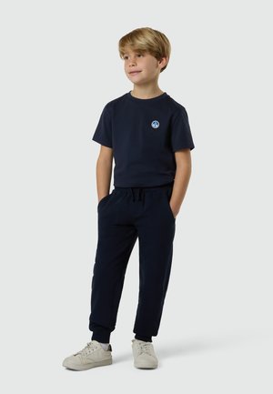 Niño de pie con las manos en los bolsillos, vistiendo una camiseta azul marino con un pequeño logo de brújula y pantalones joggers a juego, con zapatillas blancas, sobre un fondo gris.