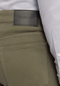 Pantalones verde oliva con un parche de cuero etiquetado "TRUSSARDI", con detalles de costura sutiles y una textura de tela suave.