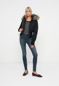 Chaqueta de plumas negra con capucha de piel sintética, llevada sobre una camiseta a rayas. Combinada con jeans oscuros de corte ajustado y mocasines burdeos brillantes.