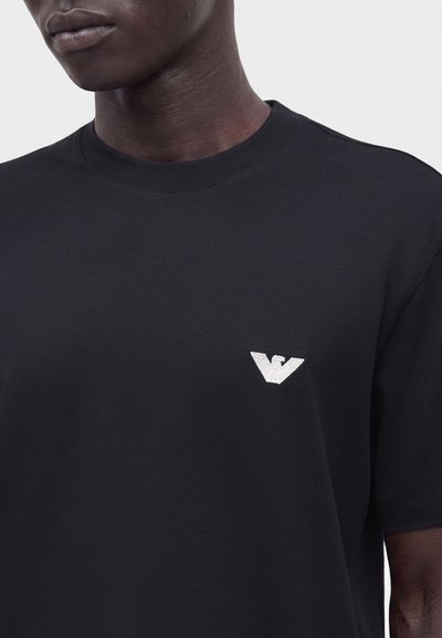 Άνδρας φοράει απλό μαύρο T-shirt με στρογγυλή λαιμόκοψη και μικρό λευκό λογότυπο αετού στο αριστερό στήθος, με φόντο μονόχρωμο.