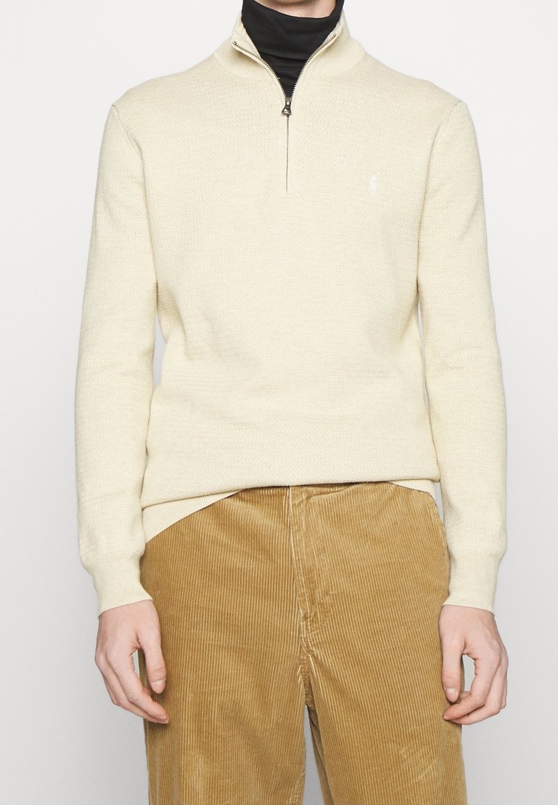 Ljus beige halvdragkedja-tröja gjort av texturerad stickad tyg, med ribbade muddärmar och en liten logotyp på bröstet, i kombination med bruna kordbyxor.