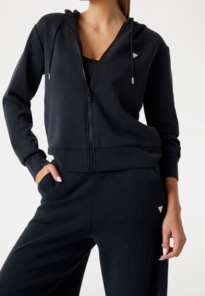 Vrouw draagt een zwarte hoodie met rits en trekkoorden en bijpassende zwarte broek, handen in de zakken, tegen een effen achtergrond.