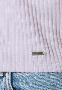 Suéter de rayas de color púrpura claro con una etiqueta de metal plateada "Hollister" en el dobladillo, combinado con jeans de mezclilla azules que exhiben una tela texturizada.