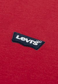 Κόκκινο βαμβακερό ύφασμα με υφή, που διαθέτει μαύρο χαρακτηριστικό Levi's με λευκά γράμματα και γραφικό σχέδιο κορώνας.