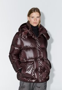 Burgundy puffer jacket with a shiny texture, hood, and two large front pockets. Features a zip closure and elastic cuffs for warmth.