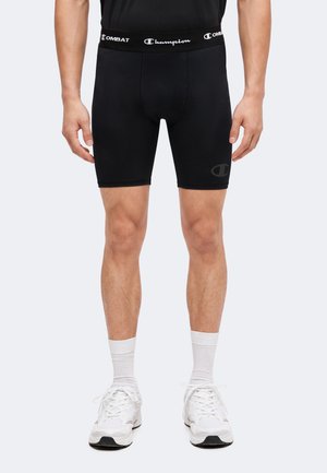 Homme portant un short de compression Champion noir, des chaussettes blanches et des chaussures de sport blanches, debout devant un fond uni.