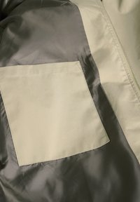 Chaqueta con un bolsillo verde oliva mate, de tejido gris brillante en contraste y una textura suave. Presenta una cremallera a lo largo del lado.