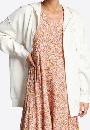 Personne portant une robe fleurie rose avec des feuilles jaunes sous un sweat à capuche blanc zippé avec le texte « la vie en rose » sur la poitrine.