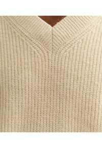 Beige gestrickter Pullover mit V-Ausschnitt, strukturierten Rippen und weichem Material, das ein warmes und komfortables Aussehen betont.
