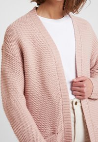 Helles rosa gestricktes Cardigan mit lockerer Passform, das ein strukturiertes Muster, offene Vorderseite und gerippte Säume aufweist. Über einem weißen Oberteil getragen.