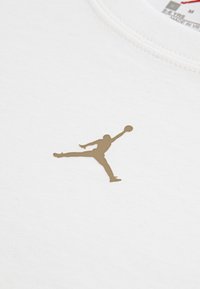 Λευκό βαμβακερό μπλουζάκι με χρυσό λογότυπο Jumpman, που απεικονίζει τη σιλουέτα ενός καλαθοσφαι ριστή σε δυναμική στάση.